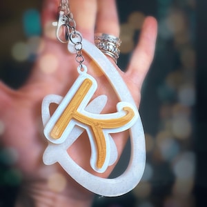 Op de afbeelding: Een witte en gouden sleutelhanger met de letter "K" in een gestileerd lettertype. De sleutelhanger is bevestigd aan een zilveren ring.