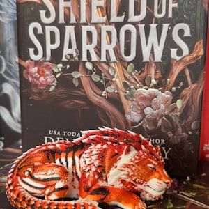 May include: A decorative tiger figurine with orange and white stripes rests in front of a book titled "Shield of Sparrows." The book cover features floral and tree branch illustrations. The figurine is a detailed, artistic representation of a tiger.