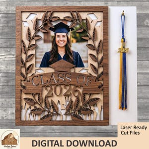 Könnte beinhalten: Ein Holz-Abschluss-Fotorahmen mit dem Text "CLASS OF 2026" und einem Foto eines Absolventen. Der Rahmen hat ein lasergeschnittenes Design mit Blättern und einem Mortarboard. Eine blaue und goldene Quaste mit einem "2026"-Anhänger hängt neben dem Rahmen.