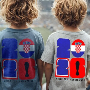 May include: Two children wearing blue and gray t-shirts with a large "2026" graphic on the back. The "2" is blue, the "0" is red, and a Croatian flag is in a circle. The text "WORLD CUP CAN MEX USA" is below.