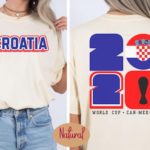 May include: Cream-colored t-shirt with the word "CROATIA" in red and blue lettering on the front. The back features the numbers "2026" in blue and red, with a Croatian flag emblem and the text "WORLD CUP CAN MEX USA".