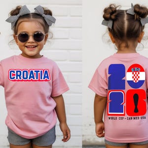 May include: A pink t-shirt with the word "CROATIA" in blue and red lettering. The back of the shirt features the numbers "2026" with the Croatian flag and text "WORLD CUP - CAN MEX USA". The child is wearing sunglasses and a bow.