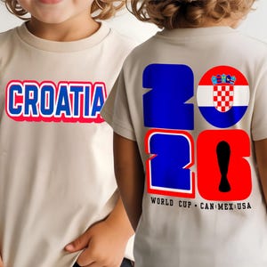 May include: Beige t-shirts with the word "CROATIA" in blue with a red outline on the front. The back of the shirt displays "2026" in blue and red, with a Croatian flag and the text "WORLD CUP CAN MEX USA".