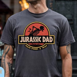 Pode incluir: T-shirt cinzenta escura com um design que diz "JURASSIC DAD" num retângulo, com um esqueleto e as palavras "PAIN FINDS A WAY" num círculo. O design é vermelho, amarelo e preto.