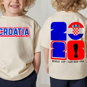 May include: A cream-colored t-shirt with the word "CROATIA" in blue and red lettering on the front. The back of the shirt features the number "2026" in blue and red, with a Croatian flag design and the text "WORLD CUP CAN MEX USA".