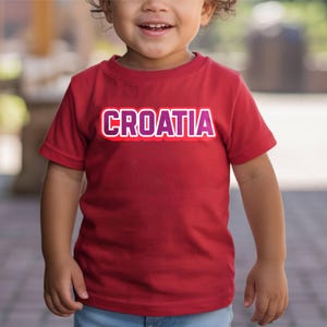 May include: A red t-shirt with the word "CROATIA" in white and purple lettering. The shirt is short-sleeved and appears to be made of cotton. The shirt is being worn by a young child.