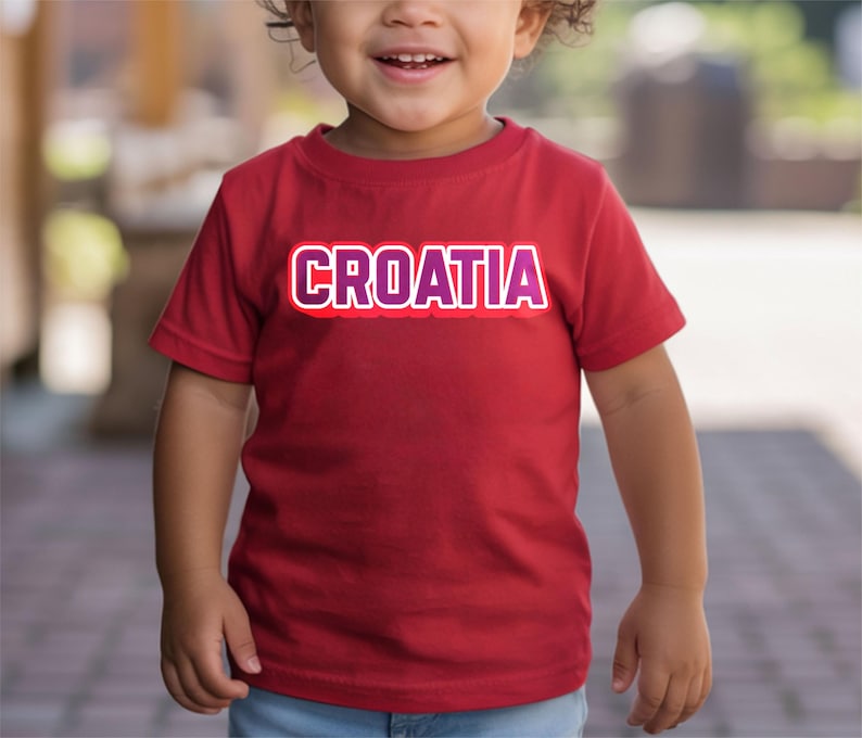 May include: A red t-shirt with the word "CROATIA" in white and purple lettering. The shirt is short-sleeved and appears to be made of cotton. The shirt is being worn by a young child.