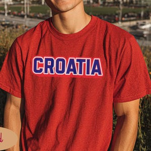 May include: A red t-shirt with the word "CROATIA" printed across the chest in white letters with a blue outline. The shirt is short-sleeved and made of a soft material. The background is blurred.