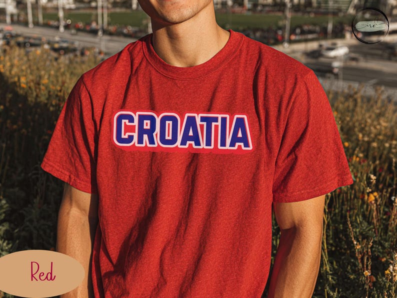 May include: A red t-shirt with the word "CROATIA" printed across the chest in white letters with a blue outline. The shirt is short-sleeved and made of a soft material. The background is blurred.