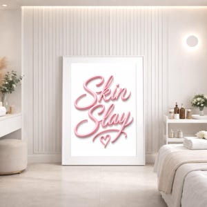 May include: A framed print with the words "Skin Slay" in pink script, with a heart symbol. The print is displayed in a white frame, leaning against a white wall. The room has a spa-like aesthetic with neutral tones.