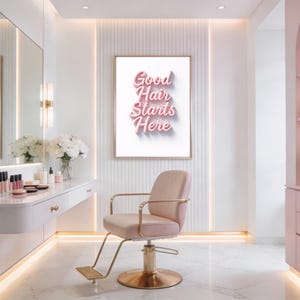 May include: A salon interior featuring a pink upholstered styling chair with a gold base, a white wall-mounted vanity with makeup brushes, and a framed wall print with the pink text Good Hair Starts Here centered on a white background.