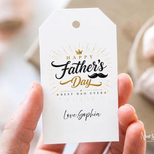 Puede incluir: Una etiqueta de regalo blanca con un diseño dorado y negro de "Happy Father's Day", un bigote y el texto "Best Dad Ever". La etiqueta también incluye el nombre "Love Sophia" en escritura cursiva.