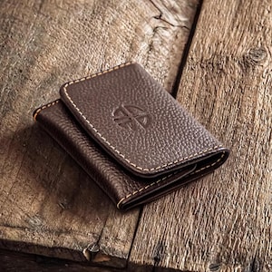 Puede incluir: Una cartera de cuero marrón con superficie texturizada y costuras blancas. La cartera tiene un emblema estampado en la parte delantera. La cartera está cerrada y parece vacía. Está sobre una superficie de madera.