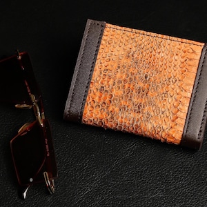 May include: A rectangular wallet featuring a textured orange snakeskin pattern on the centre panel with dark brown leather borders on the sides, accompanied by a pair of rimless sunglasses with dark brown lenses and gold-toned metal frames.
