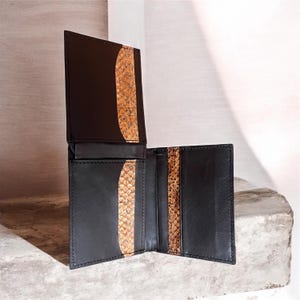 May include: A black leather bifold wallet featuring a vertical strip of textured, tan and brown snakeskin-patterned inlay on the interior panels. The wallet is open, displaying multiple card slots and a smooth, matte black finish on the leather.