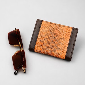 May include: A rectangular wallet featuring a central panel with a textured orange snakeskin pattern flanked by dark brown leather edges, positioned next to a pair of dark brown rectangular sunglasses with gold-tone metal hinges and accents.