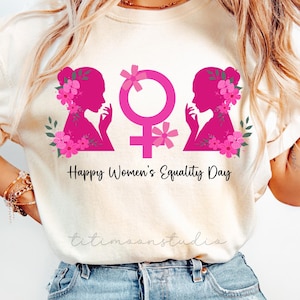 Könnte beinhalten: Cremefarbenes T-Shirt mit einem rosa Aufdruck, der zwei weibliche Silhouetten, ein Geschlechtssymbol und florale Akzente zeigt. Der Text "Happy Women's Equality Day" ist darunter gedruckt. Das Shirt ist aus weichem Material.