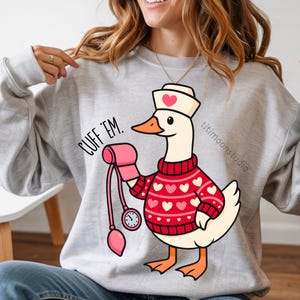 May include: A light gray sweatshirt with a cartoon goose wearing a nurse's hat and a red sweater with white hearts. The goose is holding a pink blood pressure cuff and the text "CUFF 'EM." is printed on the sweatshirt.