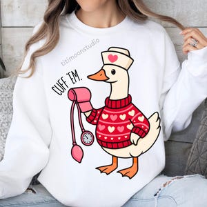 May include: White sweatshirt featuring a cartoon goose wearing a nurse's hat and a red sweater with white hearts. The goose holds a pink blood pressure cuff. Text on the shirt reads "CUFF 'EM."