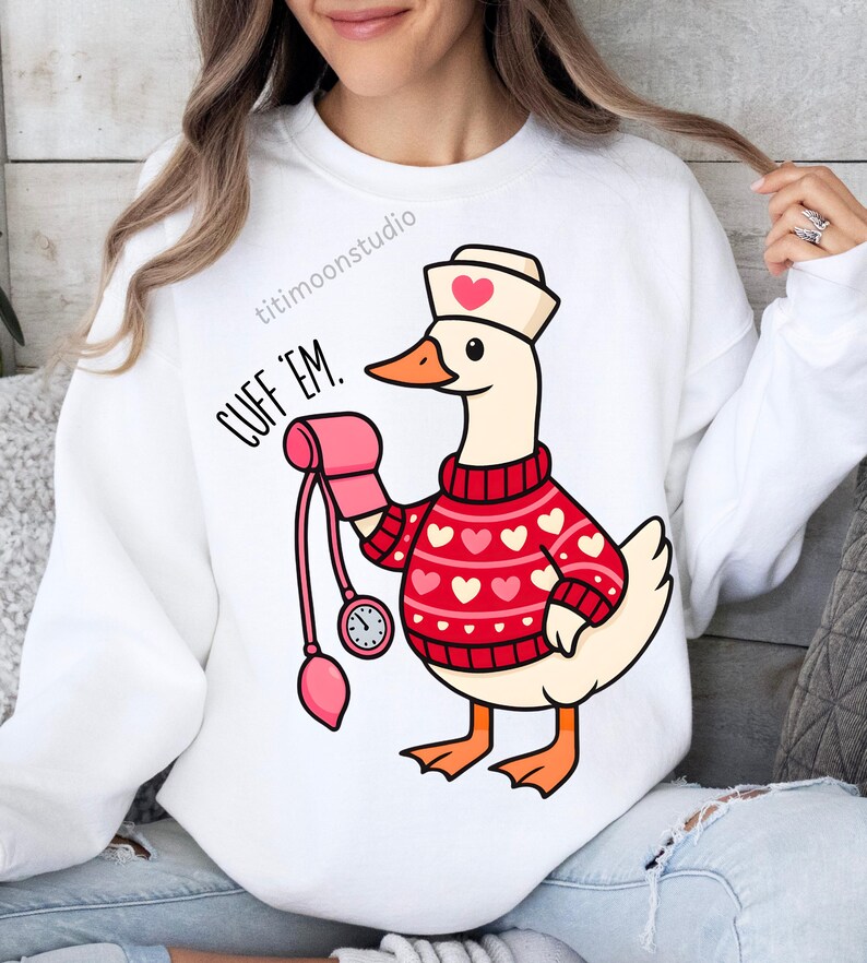 May include: White sweatshirt featuring a cartoon goose wearing a nurse's hat and a red sweater with white hearts. The goose holds a pink blood pressure cuff. Text on the shirt reads "CUFF 'EM."