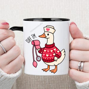 May include: White ceramic mug with a black interior and handle. The mug features a cartoon goose wearing a nurse's hat and a red sweater with white hearts. The goose is holding a pink blood pressure cuff. Text on the mug reads "CUFF 'EM."