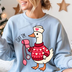 May include: Light blue sweatshirt featuring a cartoon goose dressed as a nurse, wearing a red sweater with pink hearts. The goose holds a blood pressure cuff and the text "CUFF 'EM" is printed on the left side. The sweatshirt has a crew neck.