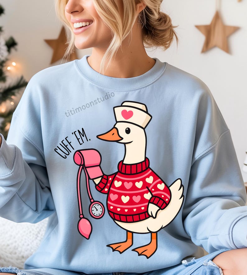 May include: Light blue sweatshirt featuring a cartoon goose dressed as a nurse, wearing a red sweater with pink hearts. The goose holds a blood pressure cuff and the text "CUFF 'EM" is printed on the left side. The sweatshirt has a crew neck.