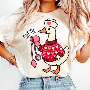 May include: Cream-colored t-shirt featuring a cartoon goose dressed as a nurse, wearing a red sweater with white hearts and a nurse's cap with a heart. The goose holds a pink blood pressure cuff. Text on the shirt reads "CUFF 'EM."