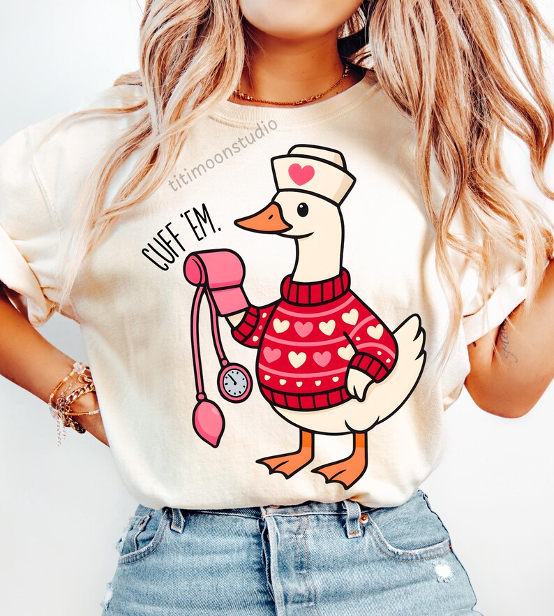 May include: Cream-colored t-shirt featuring a cartoon goose dressed as a nurse, wearing a red sweater with white hearts and a nurse's cap with a heart. The goose holds a pink blood pressure cuff. Text on the shirt reads "CUFF 'EM."