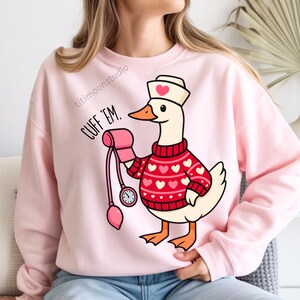 May include: Pink sweatshirt with a cartoon goose wearing a nurse's hat and a red sweater with white hearts. The goose holds a blood pressure cuff. Text on the sweatshirt reads "CUFF 'EM."