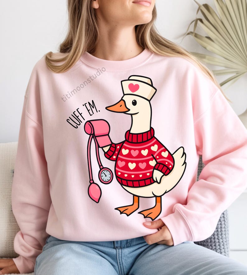 May include: Pink sweatshirt with a cartoon goose wearing a nurse's hat and a red sweater with white hearts. The goose holds a blood pressure cuff. Text on the sweatshirt reads "CUFF 'EM."