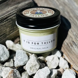 May include: A small glass jar with a black lid and a white label that reads "T is for Tallow Original Balm". The jar is sitting on a bed of small gray rocks.