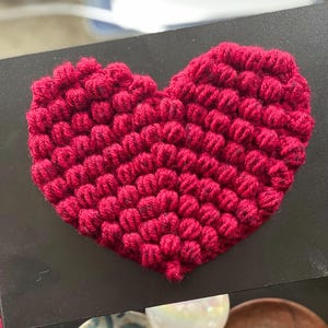 Könnte beinhalten: Ein rotes gehäkeltes Herz mit einer strukturierten, bauschigen Oberfläche. Das Herz ist aus Garn gefertigt und ein handgemachtes Handwerk.
