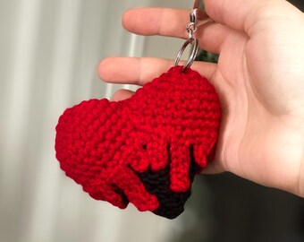 Könnte beinhalten: Ein roter gehäkelter Herz-Schlüsselanhänger mit schwarzem Sockel. Das Herz hat einen Tropfeffekt, wobei das rote Garn in die schwarze Basis zu schmelzen scheint. Der Schlüsselanhänger hat einen silberfarbenen Ring und Verschluss.
