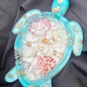 May include: A resin turtle-shaped tray with a turquoise border. The tray is filled with white sand, seashells, and pearls. The seashells are in various colors, including pink, brown, and white.