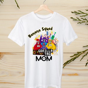 Puede incluir: Camiseta blanca con un gráfico colorido de cuatro monos tocando música. El texto "Banana Squad" está en la parte superior del gráfico y "Gorilla Tag Mom" en la parte inferior.