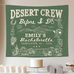 May include: Green fabric banner with white text and illustrations. The banner reads "Desert Crew Before I Do" and "Emily's Bachelorette Charleston, AZ - 2026". Illustrations include a horseshoe, ring, hat, mountains, and desert landscape.