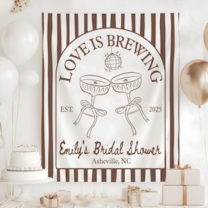 Könnte beinhalten: Ein weiß-braunes gestreiftes Banner mit dem Text "LOVE IS BREWING" und "Emily's Bridal Shower Asheville, NC". Das Banner zeigt Illustrationen von Sektgläsern und einer Discokugel. Weiße und goldene Luftballons und ein Kuchen sind im Hintergrund.