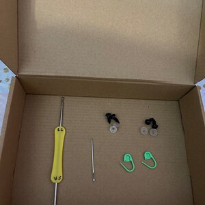 K&ouml;nnte beinhalten: Ein Karton mit einer gelben Stricknadel, einer silbernen Nadel, zwei gr&uuml;nen Sicherheitsnadeln und zwei Paaren schwarzer Kunststoff-Sicherheitsaugen mit wei&szlig;en Unterlegscheiben.