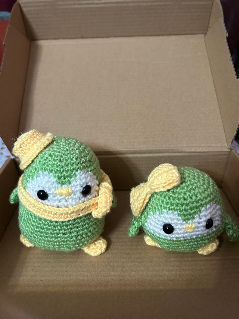 May include: Two adorable crocheted penguin plushies, one larger than the other, are displayed inside a cardboard box. The larger penguin wears a yellow hat and scarf, while the smaller one has a yellow bow. Both are crafted from soft, light green yarn with white facial features and dark eyes, creating a charming and playful aesthetic. These handmade amigurumi penguins would make delightful gifts or home decorations.