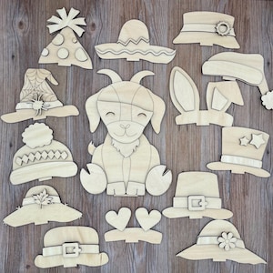 May include: A collection of wooden cutouts in various shapes, including hats, a dog, and hearts. The hats include a witch hat, a sombrero, a top hat, and a winter hat. The dog cutout is in a sitting position. The cutouts are made of light-colored wood.