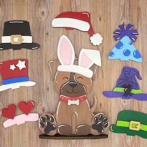 May include: Wooden cutouts of a brown dog wearing bunny ears, a Santa hat, a pilgrim hat, a patriotic hat, a witch hat, a leprechaun hat, a birthday hat, and two hearts. The dog has a red collar with a bone-shaped tag.