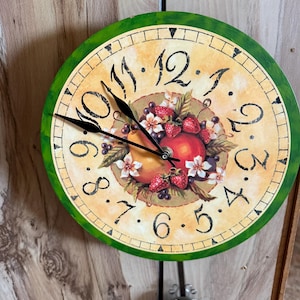 May include: Round wall clock with a green border and a cream-colored face. The clock features black numerals and hands, with a central design of fruit and flowers. A decorative pendulum hangs below.