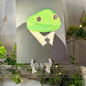 May include: A rectangular art print featuring a painted illustration of a green frog head with large dark eyes, wearing a white collared shirt and a black suit jacket. The artwork is displayed on a clear plastic stand against a dark background.