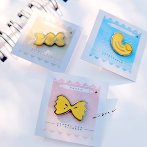 May include: Three enamel pins featuring different pasta shapes. The pins are yellow and have a glossy finish. The pins are displayed on white cards with pink, blue, and white backgrounds. The cards have the text "PASTA" and "ENAMEL PIN BY MIIBREW" printed on them.
