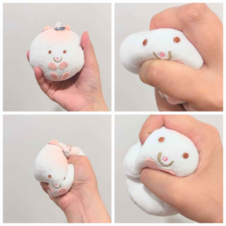 Op de afbeelding: Een wit pluche speelgoed in de vorm van een konijn met geborduurde details en roze accenten. Het speelgoed wordt in een hand geknepen, wat de zachte, squishy textuur laat zien. Het konijn heeft bruine ogen en een lachende mond.