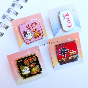 Pode incluir: Quatro pinos de esmalte com designs de embalagens de comida e bebida coreanas. Os pinos estão sobre fundos rosa e branco com o texto "Minibrew Nostalgia Pins". Os pinos representam um pacote de ramen vermelho e preto com o texto "Shin", um pacote de ramen de frango rosa e branco com texto coreano, um pacote de ramen preto e dourado com texto coreano e uma lata branca com o texto "Diet Cock".