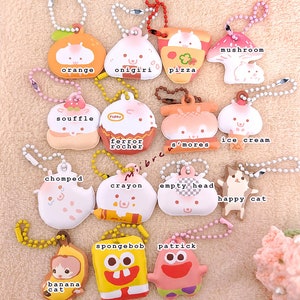 May include: A collection of 14 colorful keychains featuring various cartoon characters and food items. The keychains are made of soft, squishy material and have a variety of designs, including a mushroom, an orange, an onigiri, a pizza, a souffle, a ferrero rocher, s'mores, ice cream, a chomped character, a crayon, an empty head, a happy cat, a banana cat, Spongebob Squarepants, and Patrick Star.