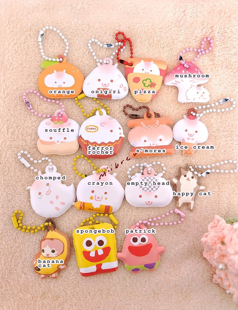 May include: A collection of 14 colorful keychains featuring various cartoon characters and food items. The keychains are made of soft, squishy material and have a variety of designs, including a mushroom, an orange, an onigiri, a pizza, a souffle, a ferrero rocher, s'mores, ice cream, a chomped character, a crayon, an empty head, a happy cat, a banana cat, Spongebob Squarepants, and Patrick Star.