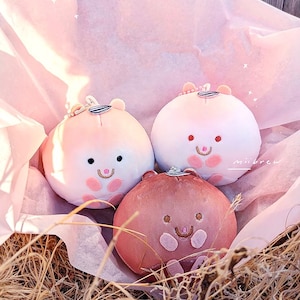 Op de afbeelding: Drie pluche, ronde, hamsterachtige speeltjes in roze en bruine tinten, genesteld in een met roze papier gevoerde mand. Elk speeltje heeft een lachend gezicht met geborduurde details en een kleine hoed. De achtergrond bevat droog gras.